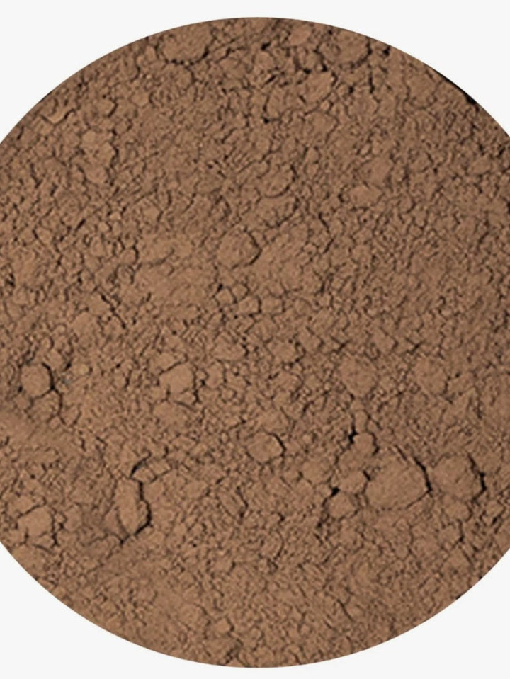 A close-up circular view of a fine brown powder or textured material filling most of the frame, with a light neutral background visible around the edges.