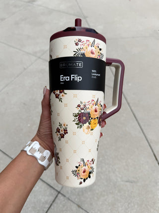 A hand holds a Brimate Era Flip 500ml insulated travel mug outdoors on a paved surface. The mug features a cream-colored body with a gold trellis pattern, a floral print, and a dark burgundy lid and handle.