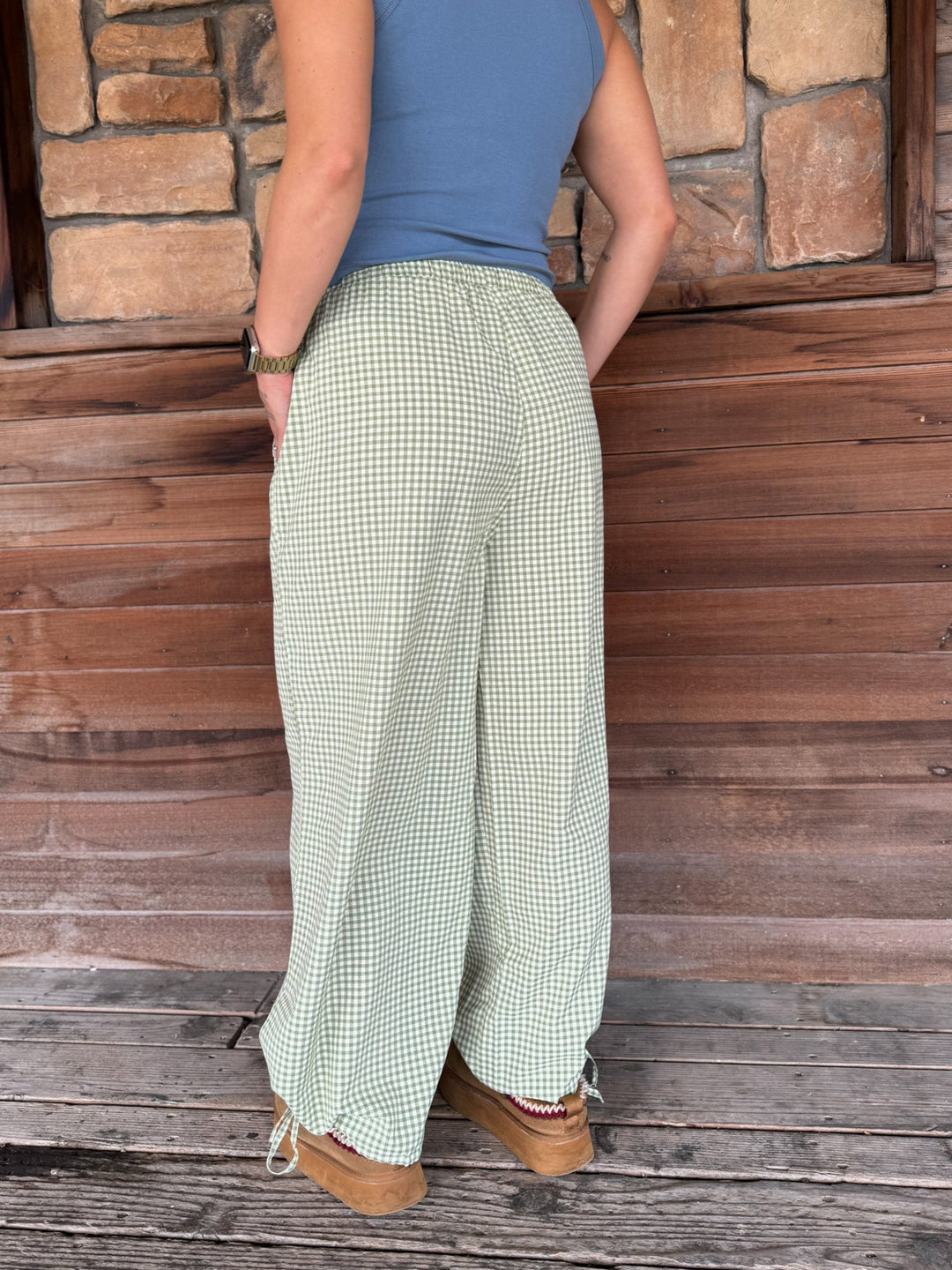 A person wearing light green and white gingham wide-leg pants, a blue sleeveless top, and tan boots, standing in front of a rustic wooden and stone wall.
