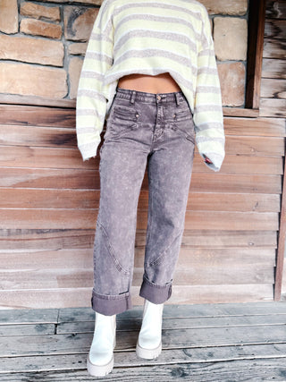 A person wearing gray acid-wash cropped jeans with rolled cuffs, a cream and gray striped cropped sweater, and white platform boots, standing against a rustic wood and stone wall.