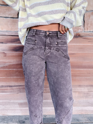 A person wearing faded brown jeans and a cream and taupe striped knit sweater, standing against a rustic wooden plank wall.