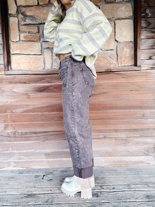 A woman stands against a rustic stone and wood wall wearing a taupe and yellow striped cropped sweater, brown wash jeans, and white platform ankle boots.