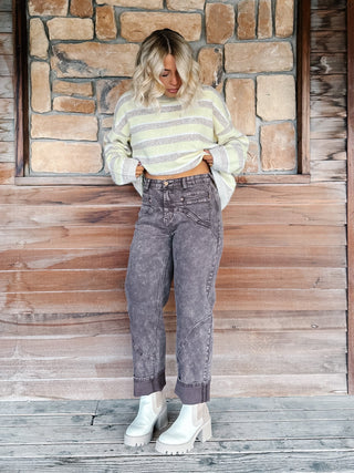A woman stands against a rustic stone and wood wall wearing a taupe and yellow striped cropped sweater, brown wash jeans, and white platform ankle boots.