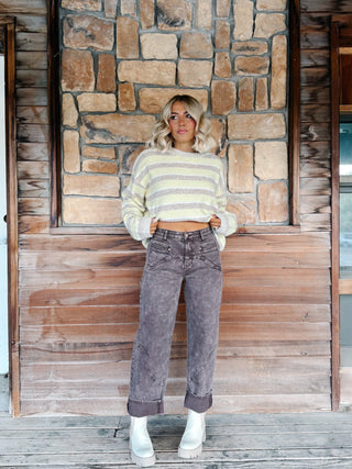 A woman stands against a rustic stone and wood wall wearing a taupe and yellow striped cropped sweater, brown wash jeans, and white platform ankle boots.