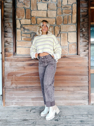 A woman stands against a rustic stone and wood wall wearing a taupe and yellow striped cropped sweater, brown wash jeans, and white platform ankle boots.