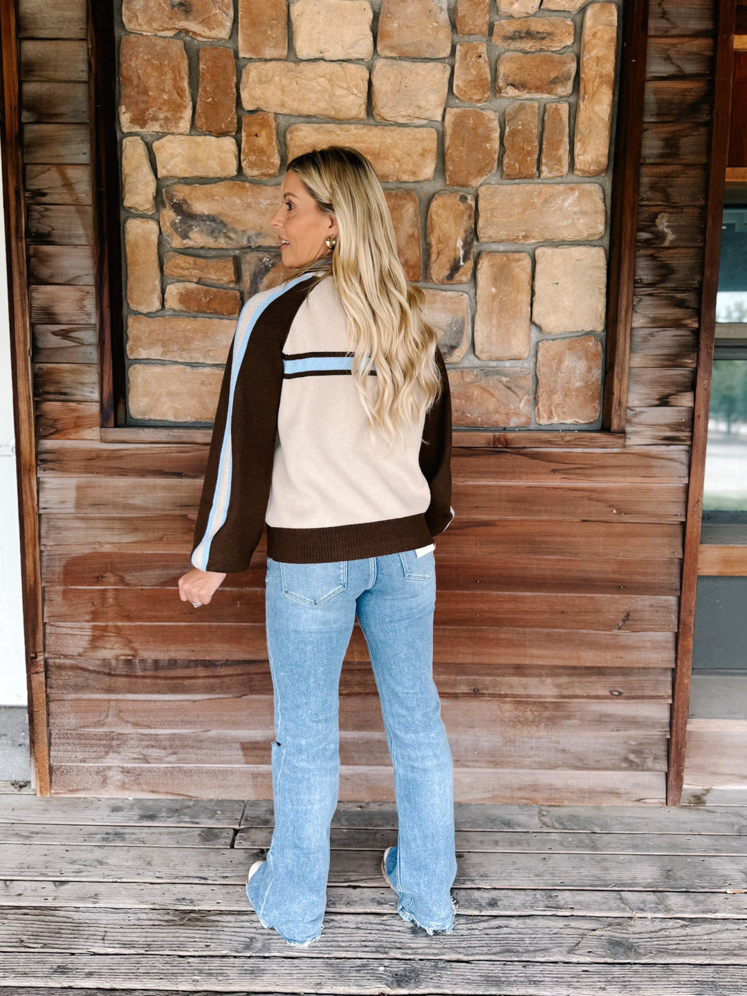A woman stands on a wooden porch in front of a stone wall, wearing a beige, brown, and light blue color-blocked track jacket with light-wash blue flare jeans.