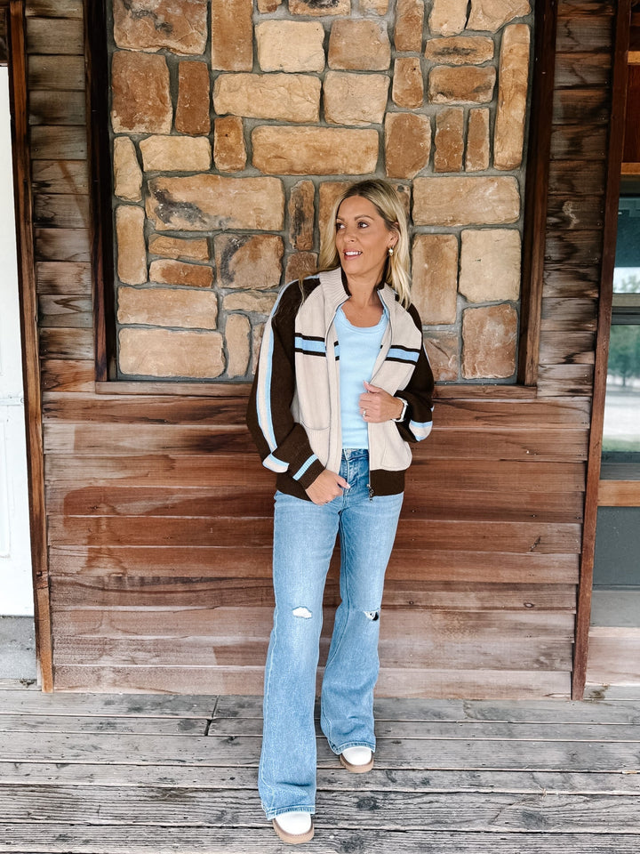 A woman stands on a wooden porch in front of a stone wall, wearing a cream, brown, and navy striped bomber jacket over a light blue top, paired with light-wash distressed jeans and white sneakers.