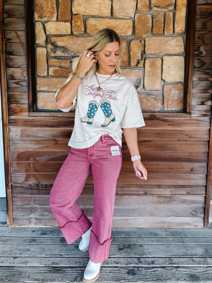 A woman posing in front of a rustic wooden and stone wall, wearing a cream graphic t-shirt with &