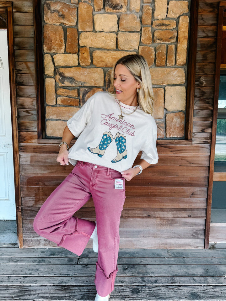 A woman posing on a rustic wooden porch in front of a stone wall, wearing a cream graphic t-shirt that reads &