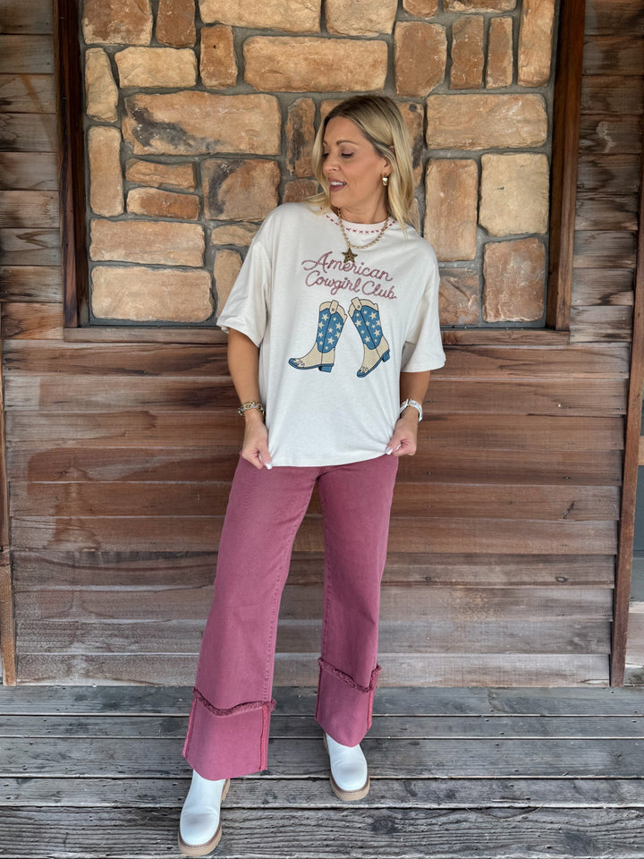 A woman stands on a wooden porch in front of a stone wall, wearing a cream graphic t-shirt that reads &