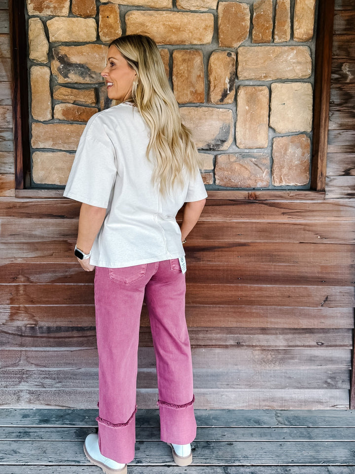 A woman stands with her back to the camera wearing a white short-sleeve top and pink wide-leg jeans with rolled cuffs, posed in front of a rustic stone and wood wall.
