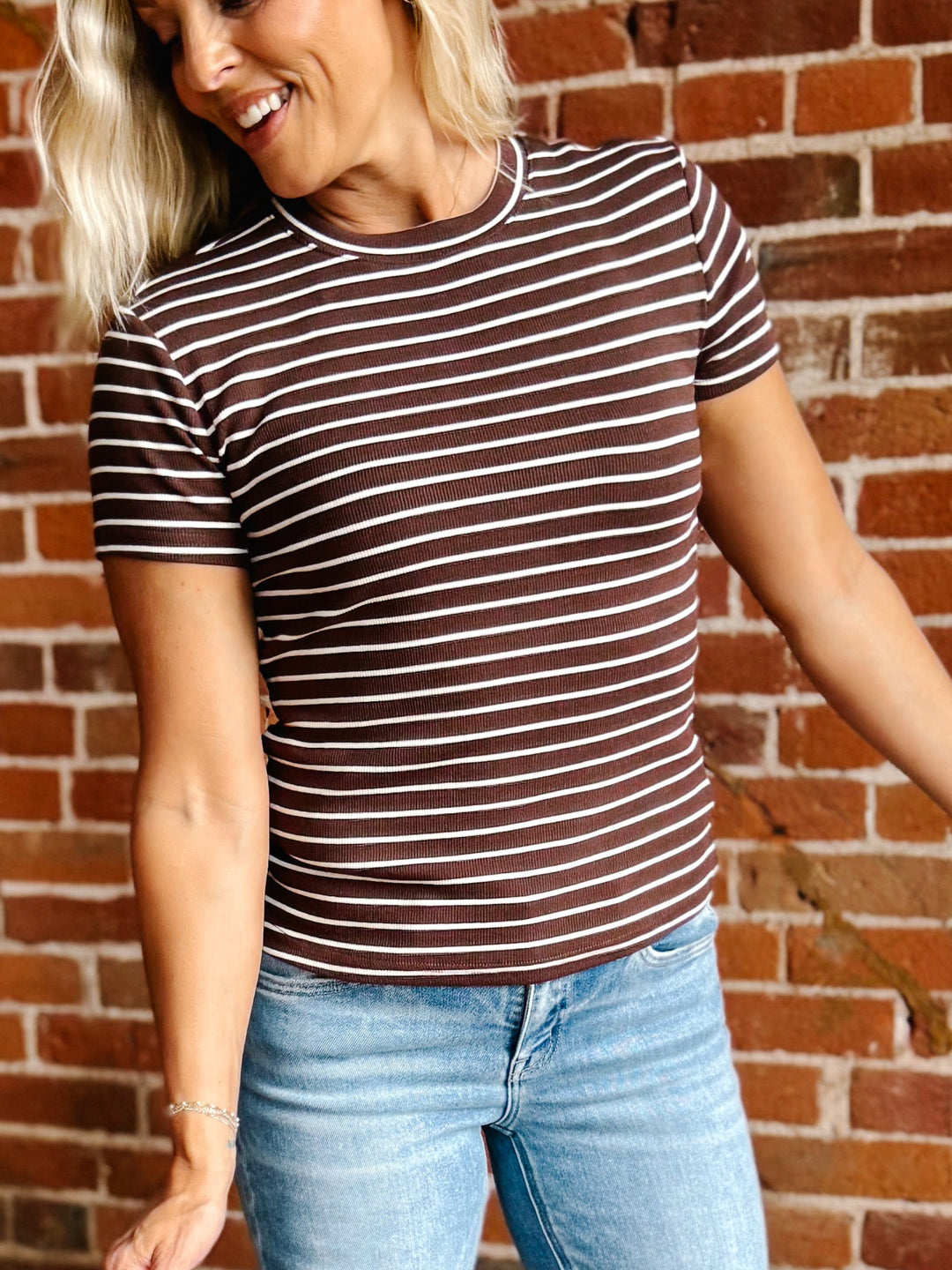 A woman wearing a brown and white horizontally striped short-sleeve t-shirt and light blue jeans, standing in front of a red brick wall.