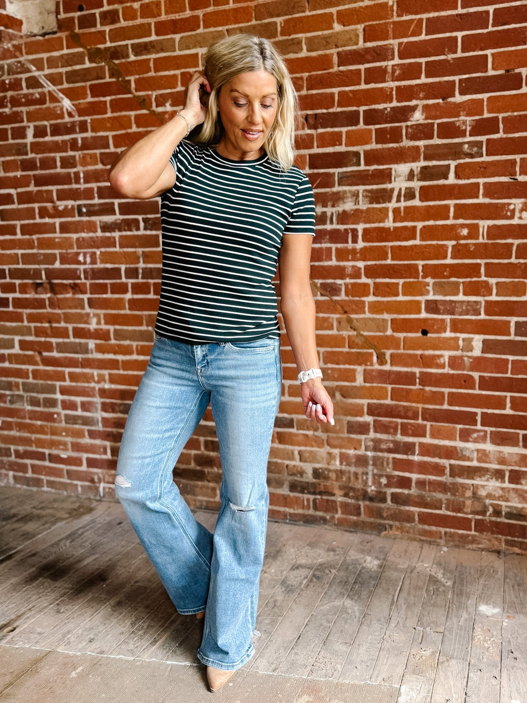 A woman with blonde hair wearing a Hunter Green and white horizontally striped t-shirt and light blue jeans, standing in front of a red brick wall.