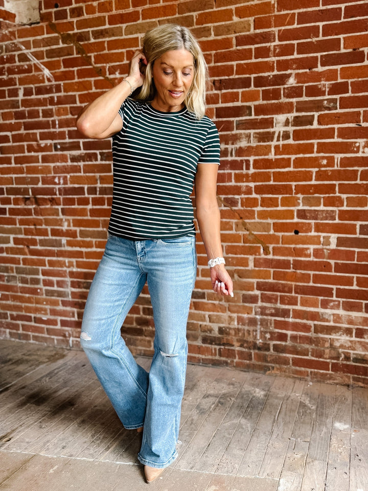 A woman with blonde hair wearing a Hunter Green and white horizontally striped t-shirt and light blue jeans, standing in front of a red brick wall.