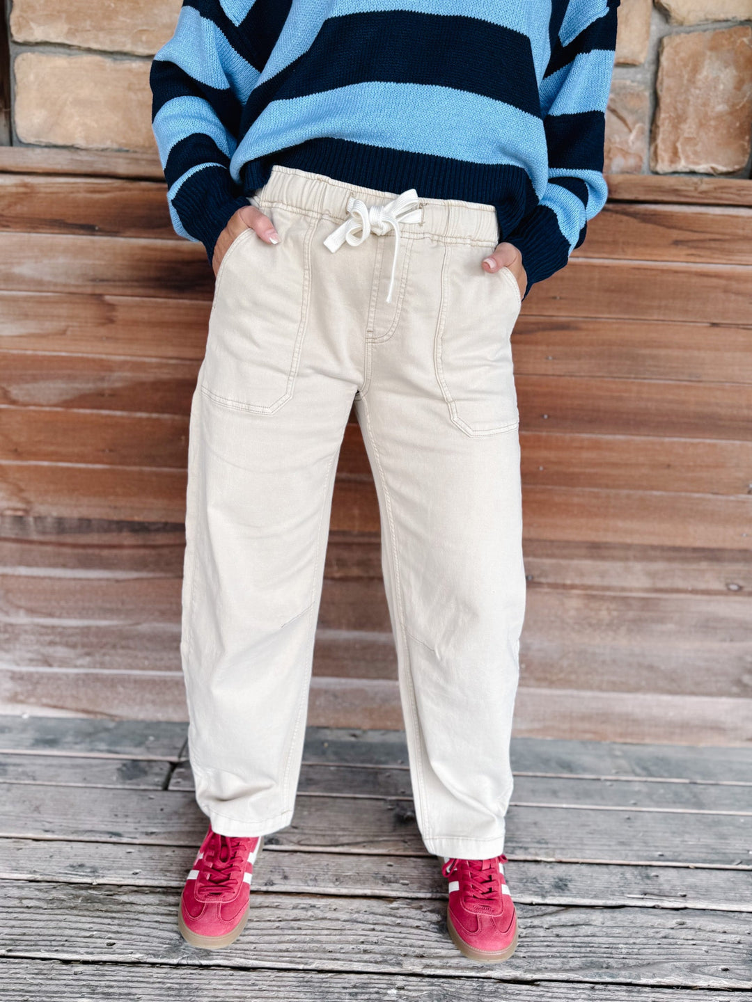 A person wearing cream-colored drawstring pants, a blue and navy striped sweater, and red sneakers, standing against a rustic wooden wall and deck.