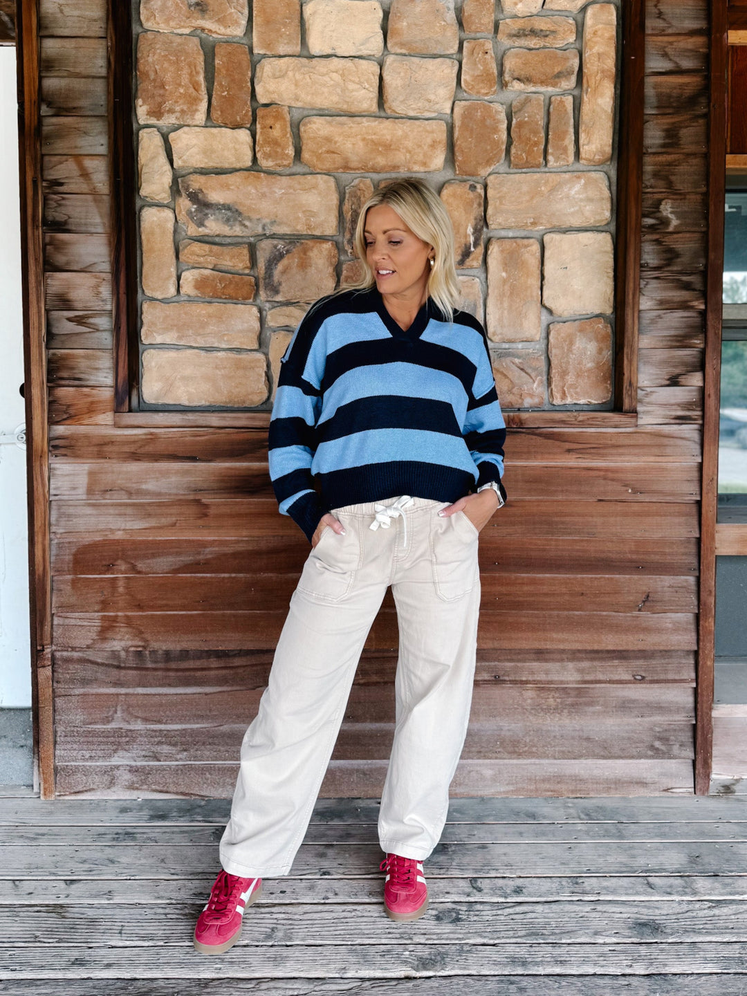 A woman stands on a wooden porch in front of a stone and wood wall, wearing a blue and black striped long-sleeve knit sweater, cream wide-leg pants, and bright pink sneakers.