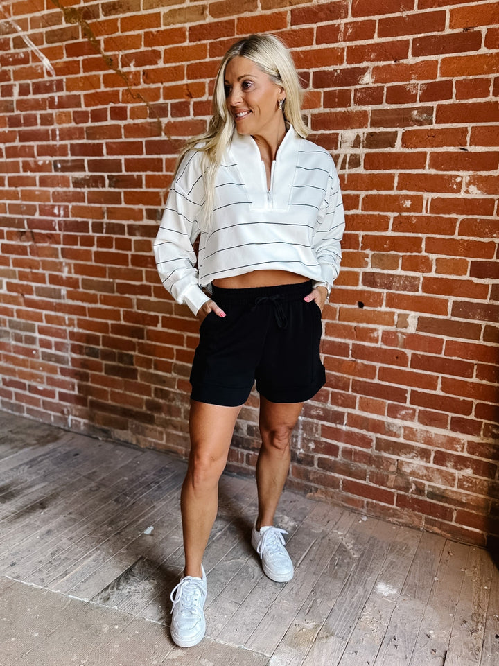 A woman stands in front of a red brick wall wearing a white half-zip pullover with thin gray horizontal stripes, black shorts, and white sneakers.