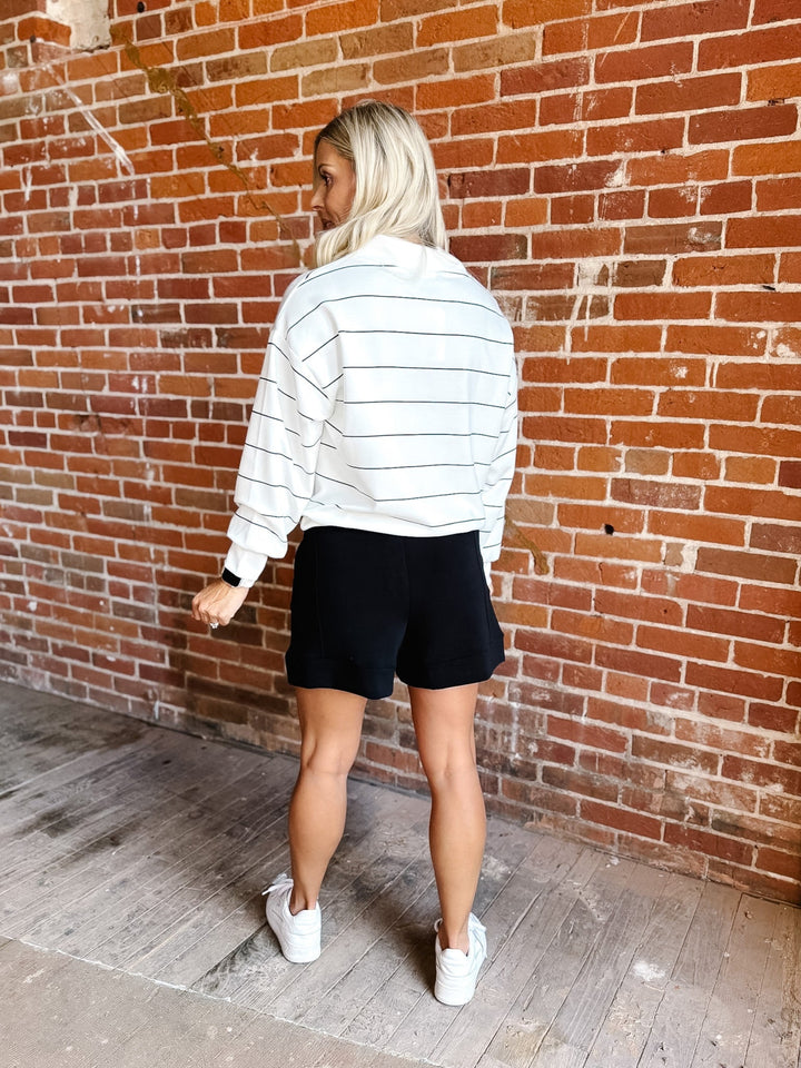 A woman standing with her back to the camera, wearing a white cropped sweatshirt with thin black horizontal stripes, black shorts, and white sneakers, set against a red brick wall background.