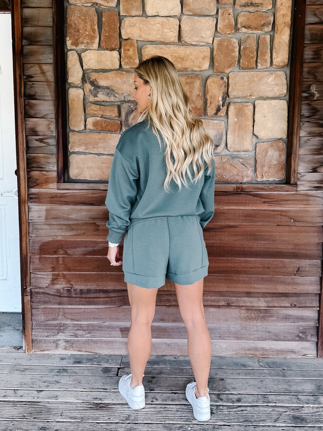 A woman stands on a rustic wooden porch wearing a sage green two-piece lounge set consisting of a long-sleeve sweatshirt and matching shorts, paired with white sneakers.