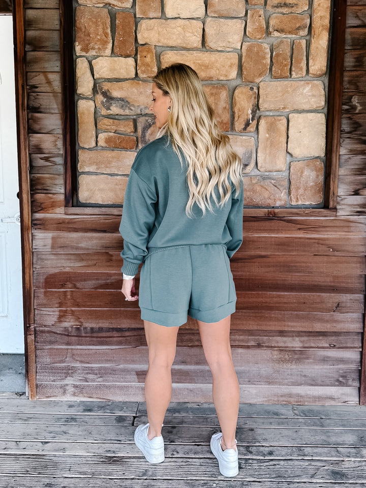 A woman stands on a rustic wooden porch wearing a sage green two-piece lounge set consisting of a long-sleeve sweatshirt and matching shorts, paired with white sneakers.
