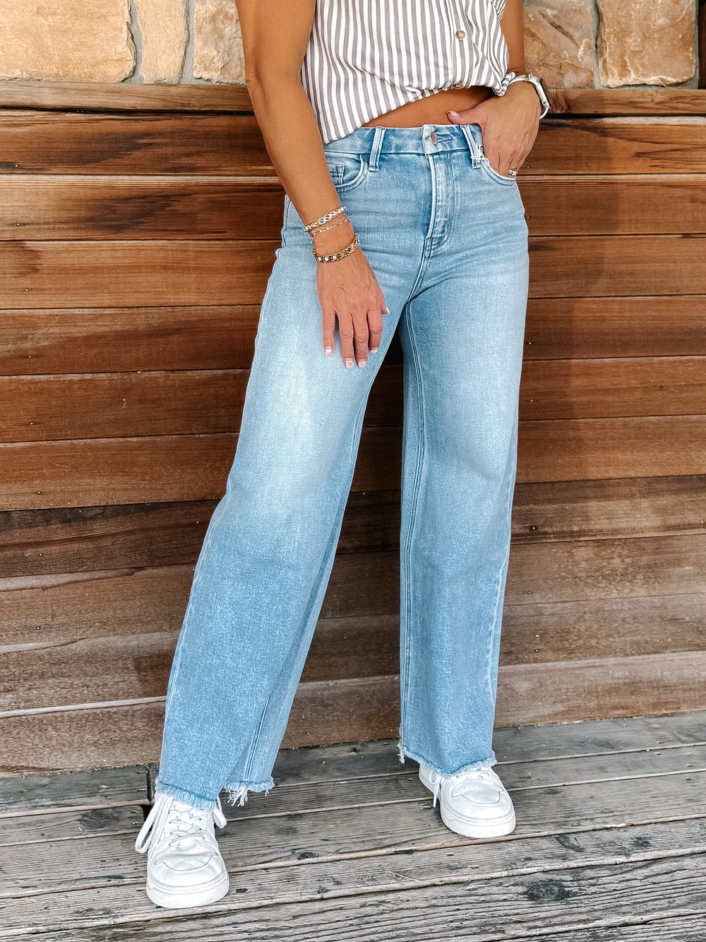 A person wearing light-wash wide-leg denim jeans with a striped crop top and white sneakers, standing against a rustic wooden wall.