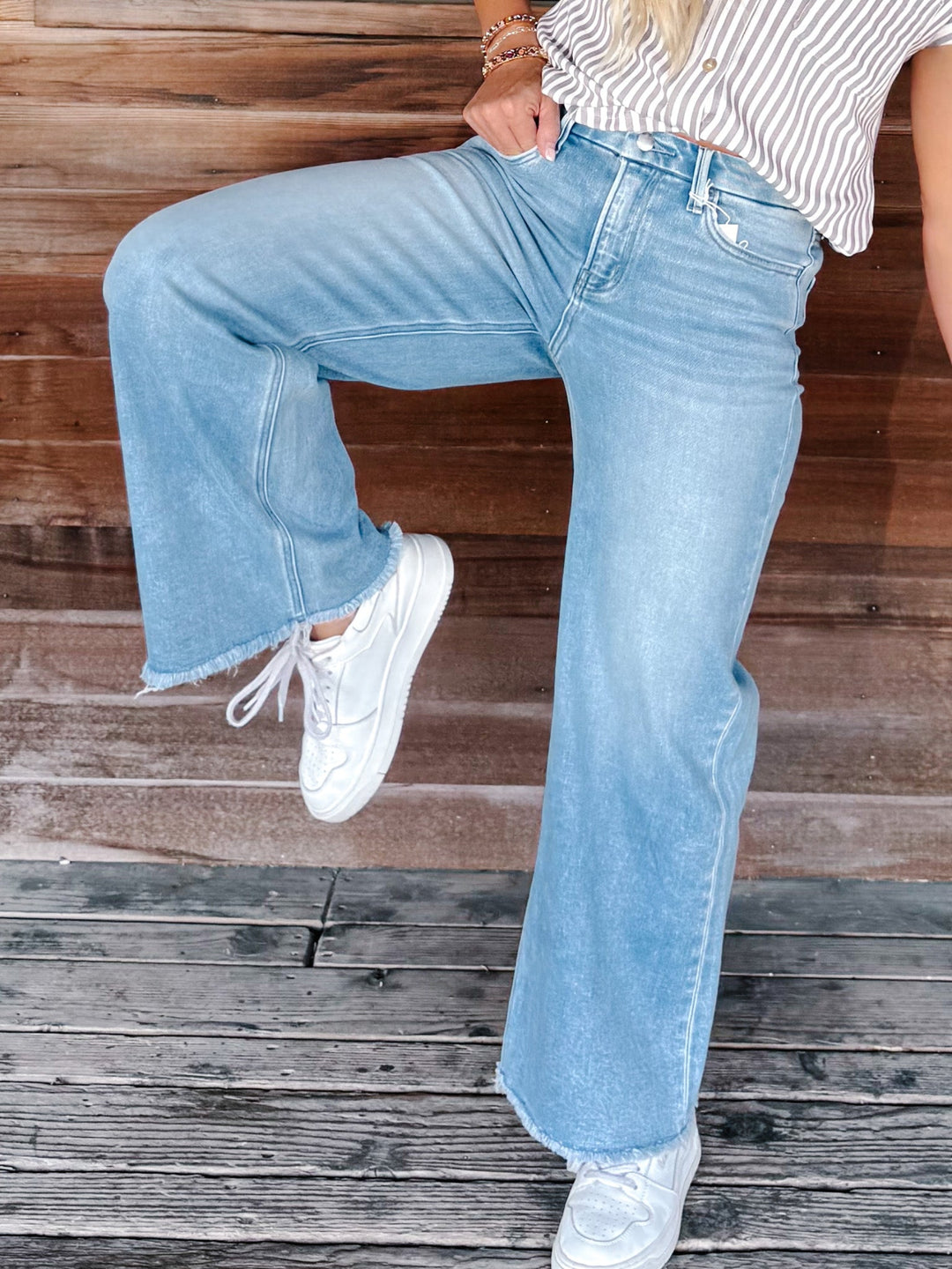 A person wearing light-wash blue wide-leg jeans with frayed hems, paired with a white and brown striped top and white sneakers, standing against a rustic wooden plank background.