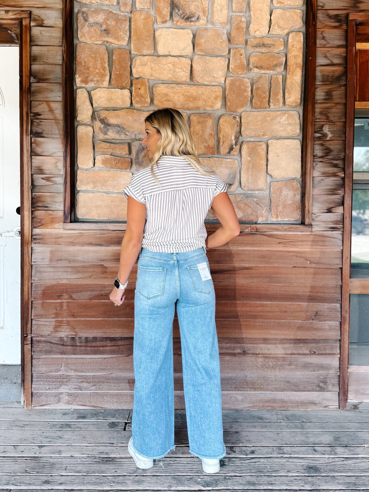 A woman stands with her back to the camera wearing a white and brown  striped short-sleeve shirt and light blue wide-leg jeans, posing in front of a rustic wooden and stone wall.