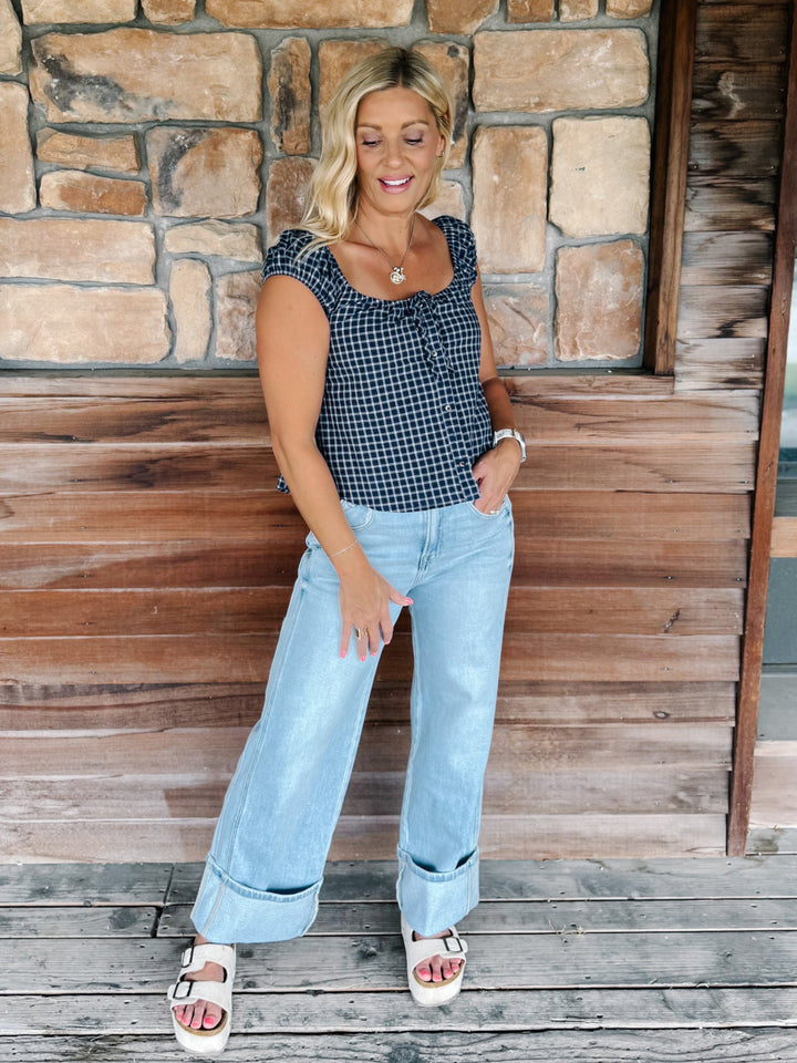A woman stands on a wooden deck in front of a stone wall, wearing a navy and white gingham sleeveless top, light blue wide-leg jeans, and white platform sandals.