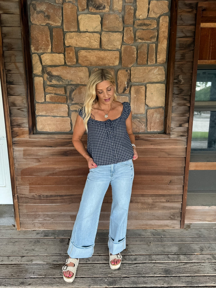 A woman stands on a wooden porch in front of a stone and wood wall, wearing a navy and white gingham sleeveless top, light blue wide-leg jeans, and beige slide sandals.