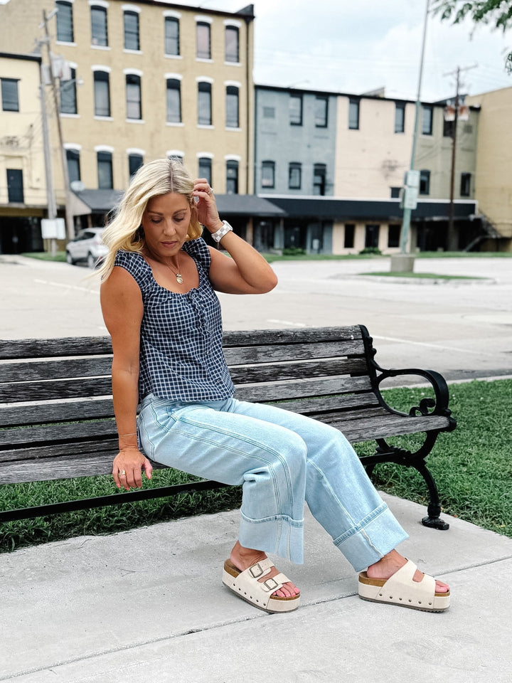 A woman sits on a wooden park bench wearing a black and white gingham sleeveless top, light blue wide-leg jeans, and beige slide sandals, with an urban street scene in the background.