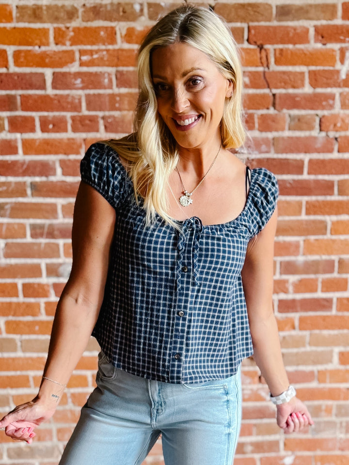 A woman wearing a navy plaid check top with a scoop neckline, button front, and tie detail.
