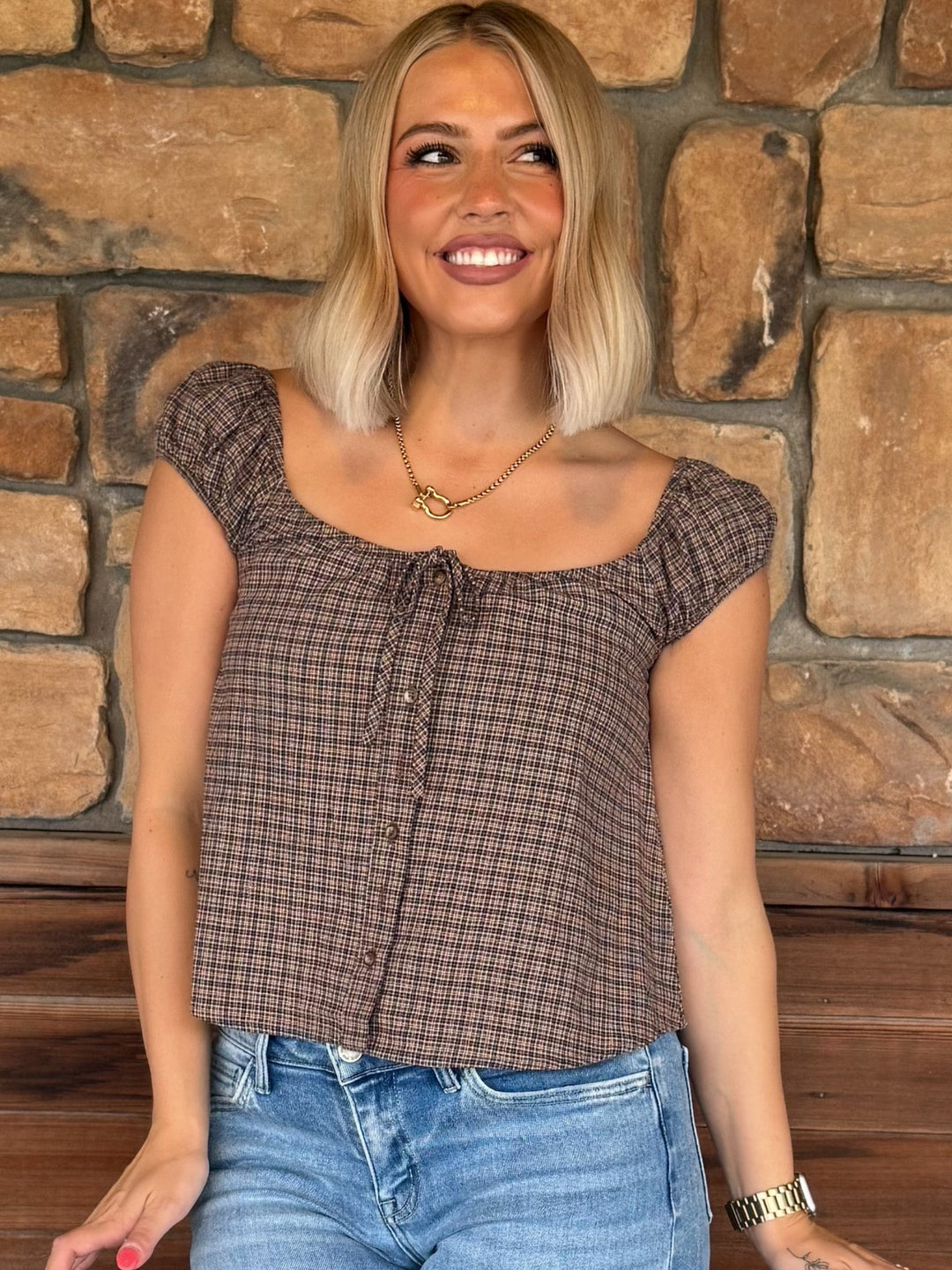 A smiling blonde woman wearing a brown and black plaid off-the-shoulder top with a tie neckline and blue jeans, standing in front of a rustic stone wall.