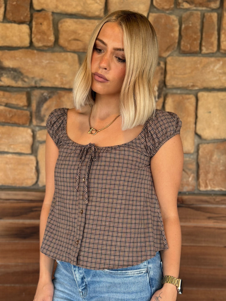 A woman with blonde hair wearing a brown and black plaid short-sleeve top with a ruffled neckline, paired with blue jeans, standing in front of a rustic stone wall.