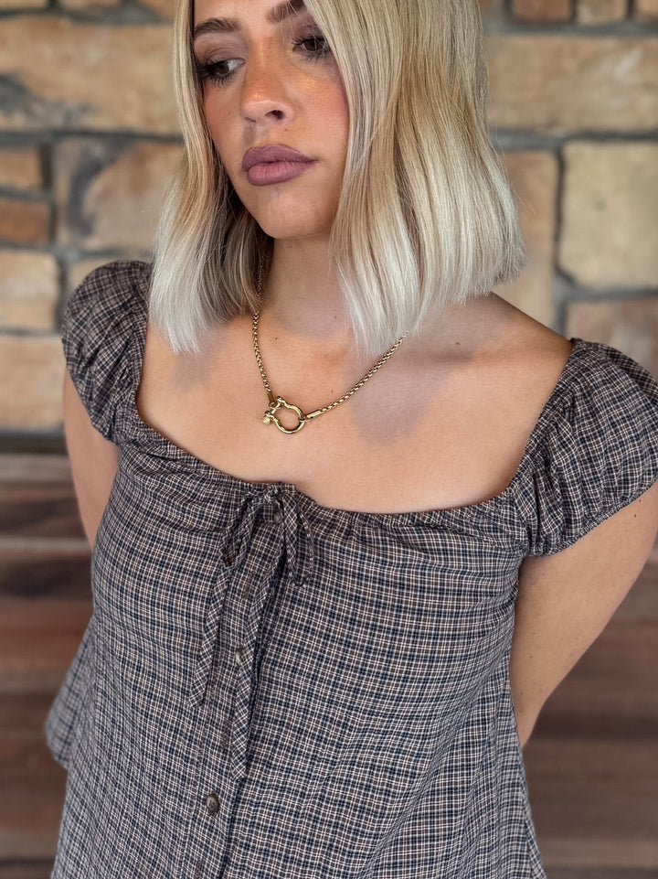 A woman with blonde hair wearing a dark gray and black plaid off-the-shoulder top with a front tie detail, paired with a gold chain necklace, set against a rustic stone wall background.