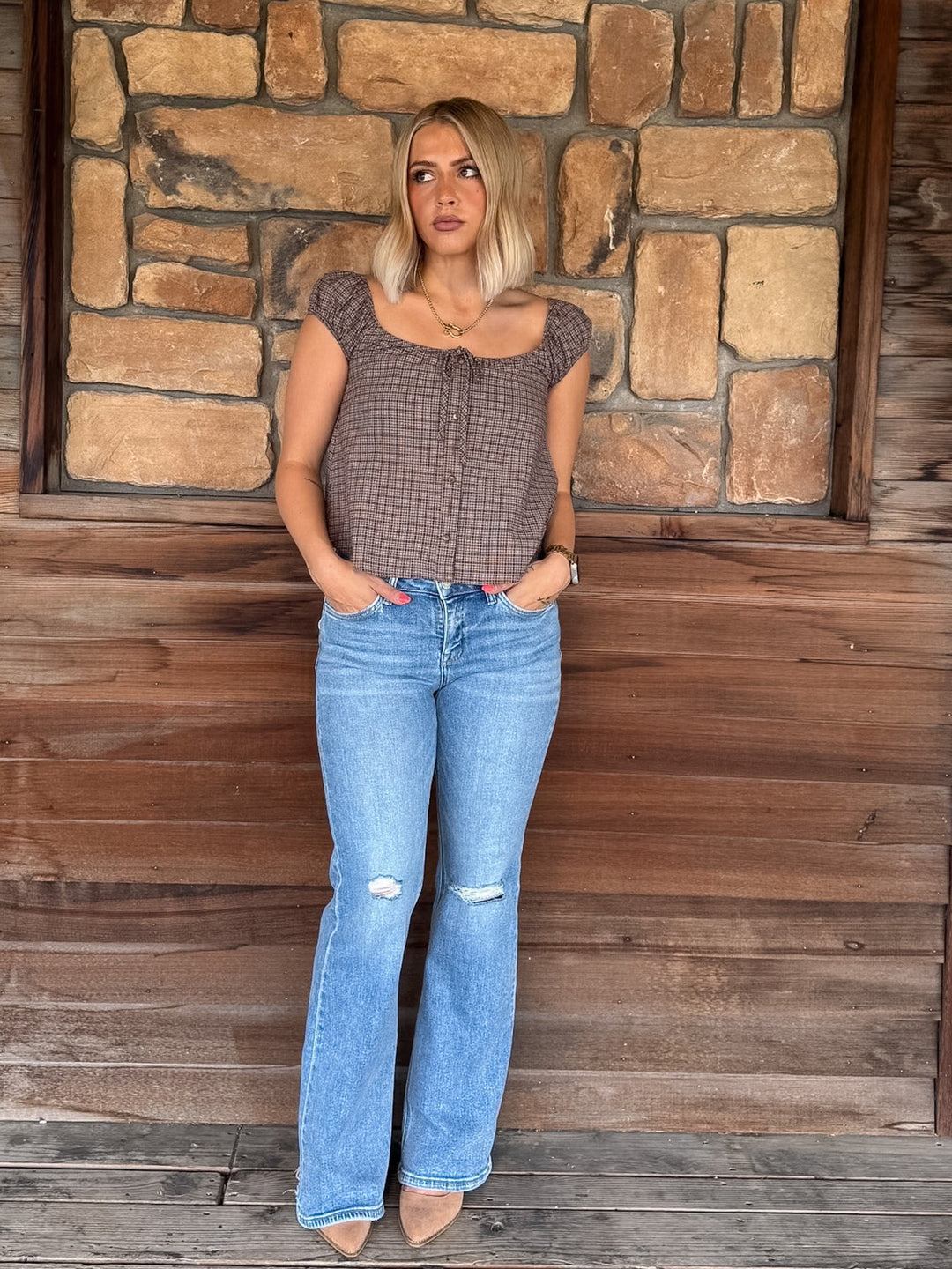 A woman stands against a rustic stone and wood wall wearing a brown and black gingham patterned crop top with cap sleeves, paired with light blue distressed flare jeans and beige heels.