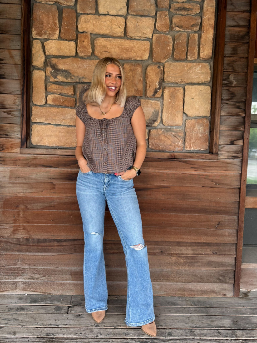 A woman stands on a wooden porch in front of a stone wall, wearing a brown and beige plaid short-sleeve top with tie details at the neckline, light-wash blue distressed flare jeans, and beige pointed-toe heels.