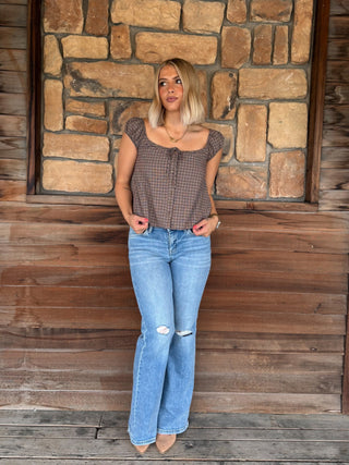 A woman stands on a rustic wooden porch in front of a stone wall, wearing a brown and black checkered short-sleeve top with a tie neckline and light-wash blue distressed flare jeans.