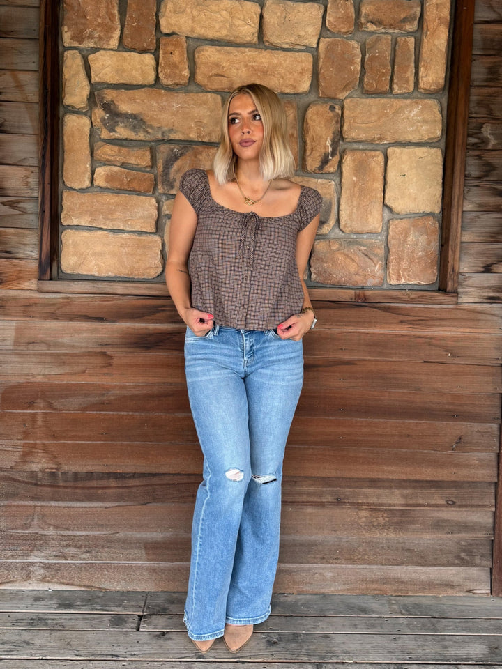 A woman stands on a rustic wooden porch in front of a stone wall, wearing a brown and black checkered short-sleeve top with a tie neckline and light-wash blue distressed flare jeans.