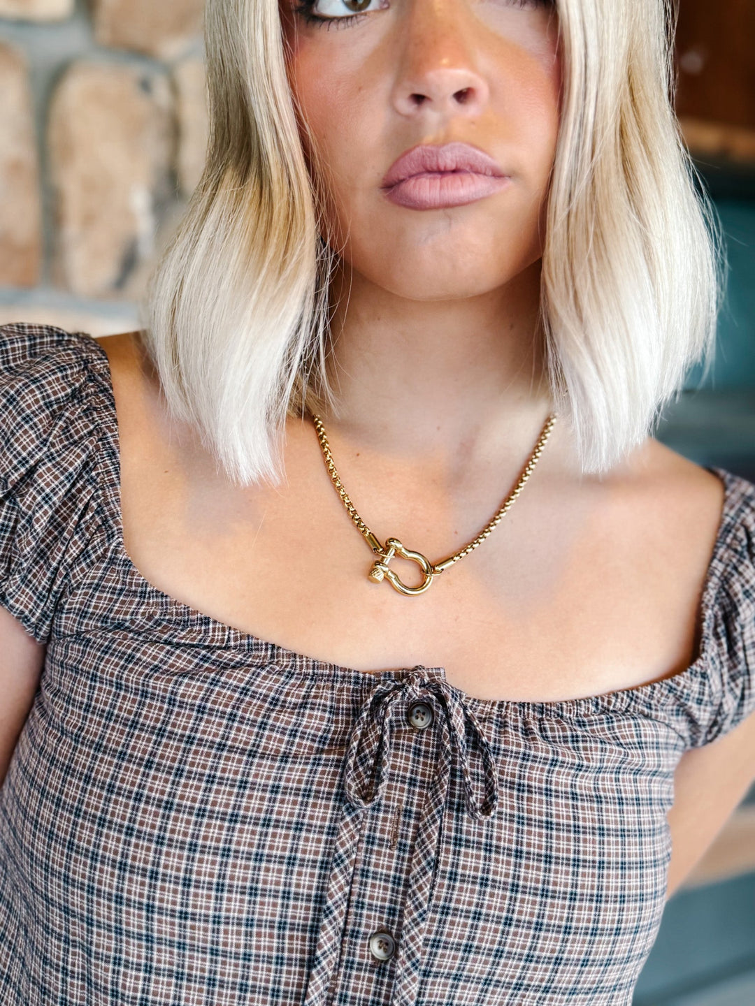 A close-up lifestyle shot of a woman with blonde hair wearing a gold chain necklace with a horse-head pendant, paired with a plaid top.