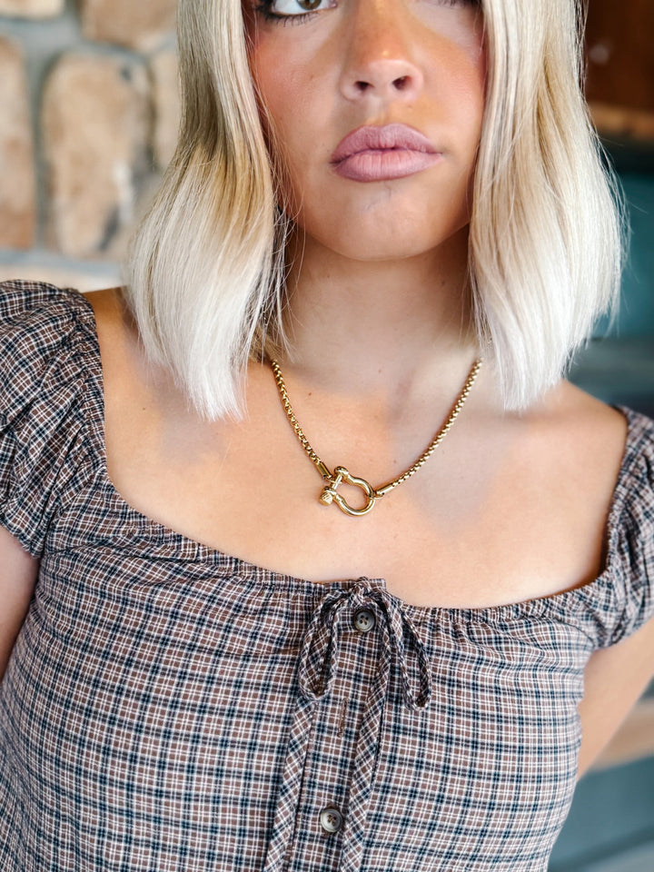 A close-up lifestyle shot of a woman with blonde hair wearing a gold chain necklace with a horse-head pendant, paired with a plaid top.