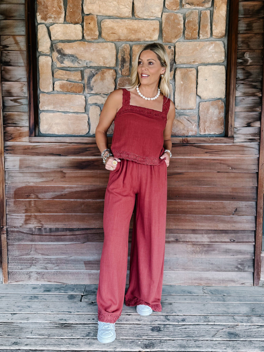 A woman modeling a rust-red two-piece linen set consisting of a sleeveless top and matching wide-leg pants, standing on a wooden porch with a stone wall background.