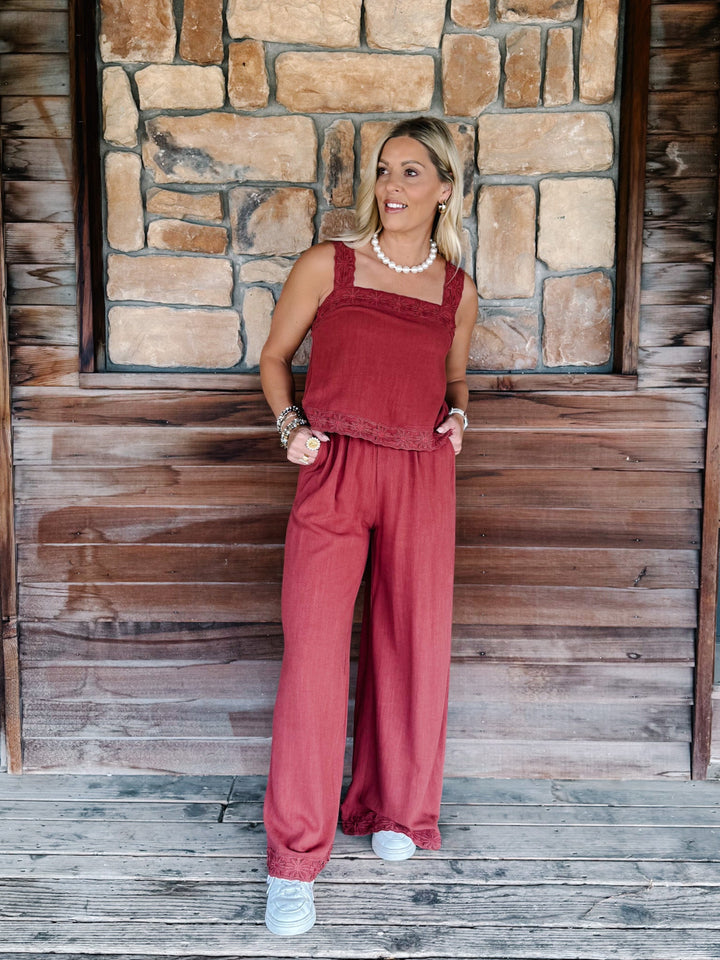 A woman modeling a rust-red two-piece linen set consisting of a sleeveless top and matching wide-leg pants, standing on a wooden porch with a stone wall background.