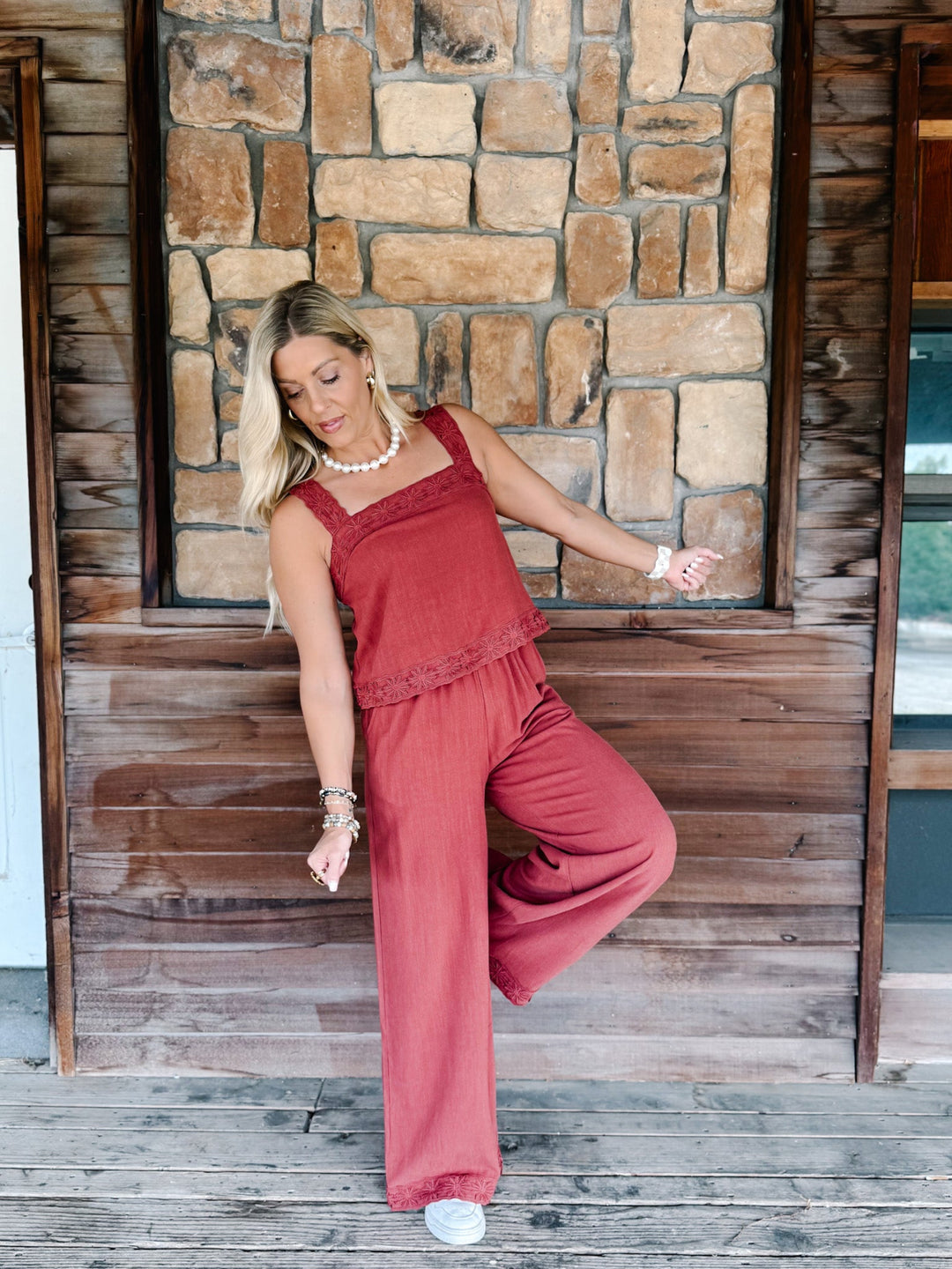 A woman modeling a rust-red two-piece linen set consisting of a ruffled camisole top and matching wide-leg pants, posing on a rustic wooden porch with a stone wall background.