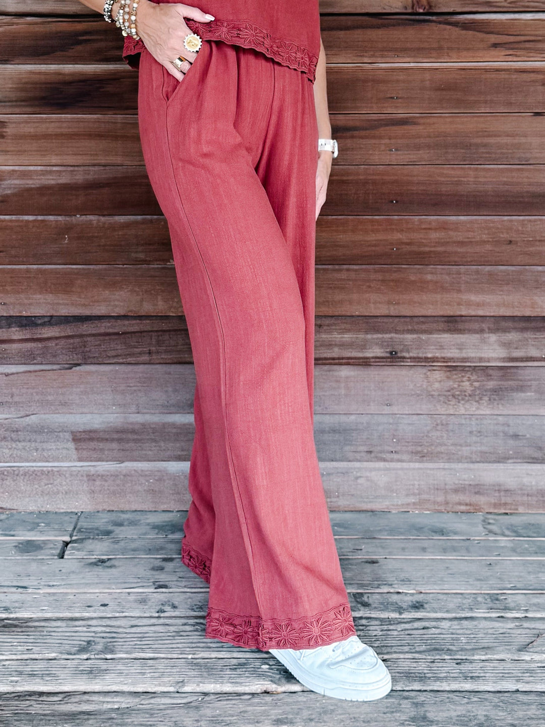 A person wearing wide-leg dusty rose pants with lace trim at the hem, paired with white sneakers, standing against a rustic wooden plank wall.