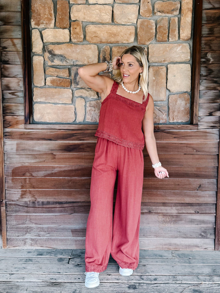 A woman modeling a dusty rose two-piece linen set consisting of a sleeveless top with lace trim and matching wide-leg pants, standing against a rustic stone and wood wall.