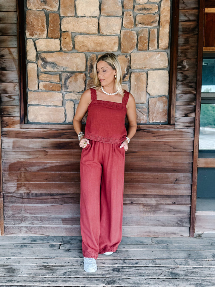 A woman stands on a wooden porch in front of a stone wall, wearing a rust-red sleeveless jumpsuit with wide legs and white sneakers.