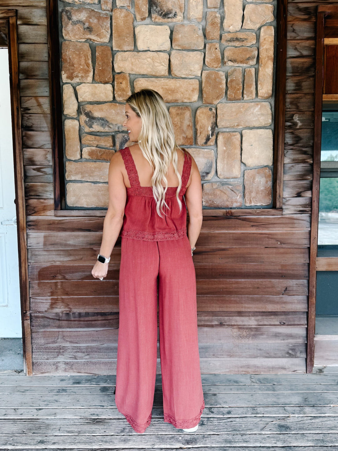 A woman stands on a wooden porch wearing a dusty rose wide-leg jumpsuit, photographed from behind against a rustic stone and wood wall.