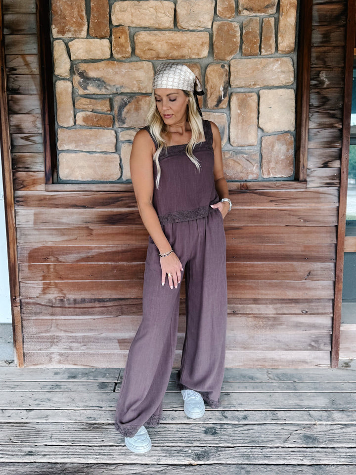 A woman wearing a mauve-purple sleeveless wide-leg jumpsuit, a beige patterned headscarf, and white sneakers, standing on a rustic wooden porch in front of a stone wall.
