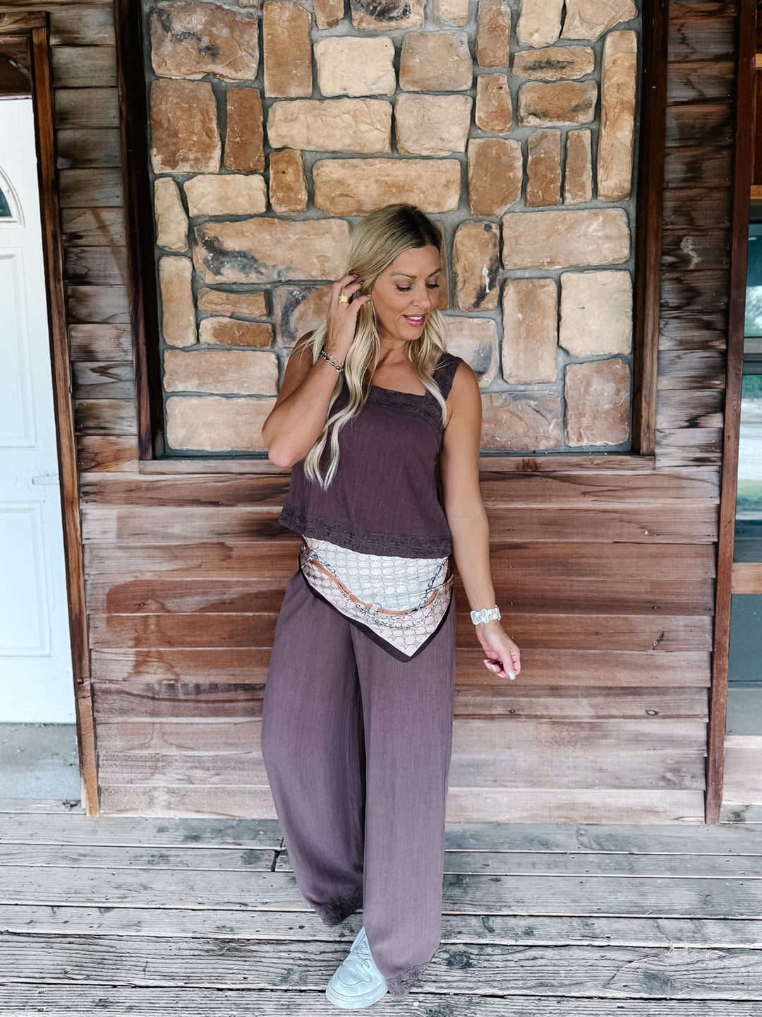A woman modeling a two-piece lounge set consisting of a sleeveless dark plum top and matching wide-leg pants, styled with a patterned bandana belt and white sneakers, standing on a wooden porch in front of a stone wall.