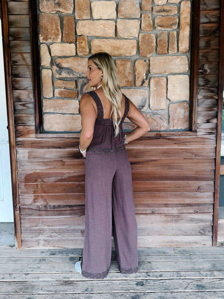 A woman stands on a wooden porch wearing a mauve wide-leg jumpsuit, photographed from the back against a rustic stone and wood wall.
